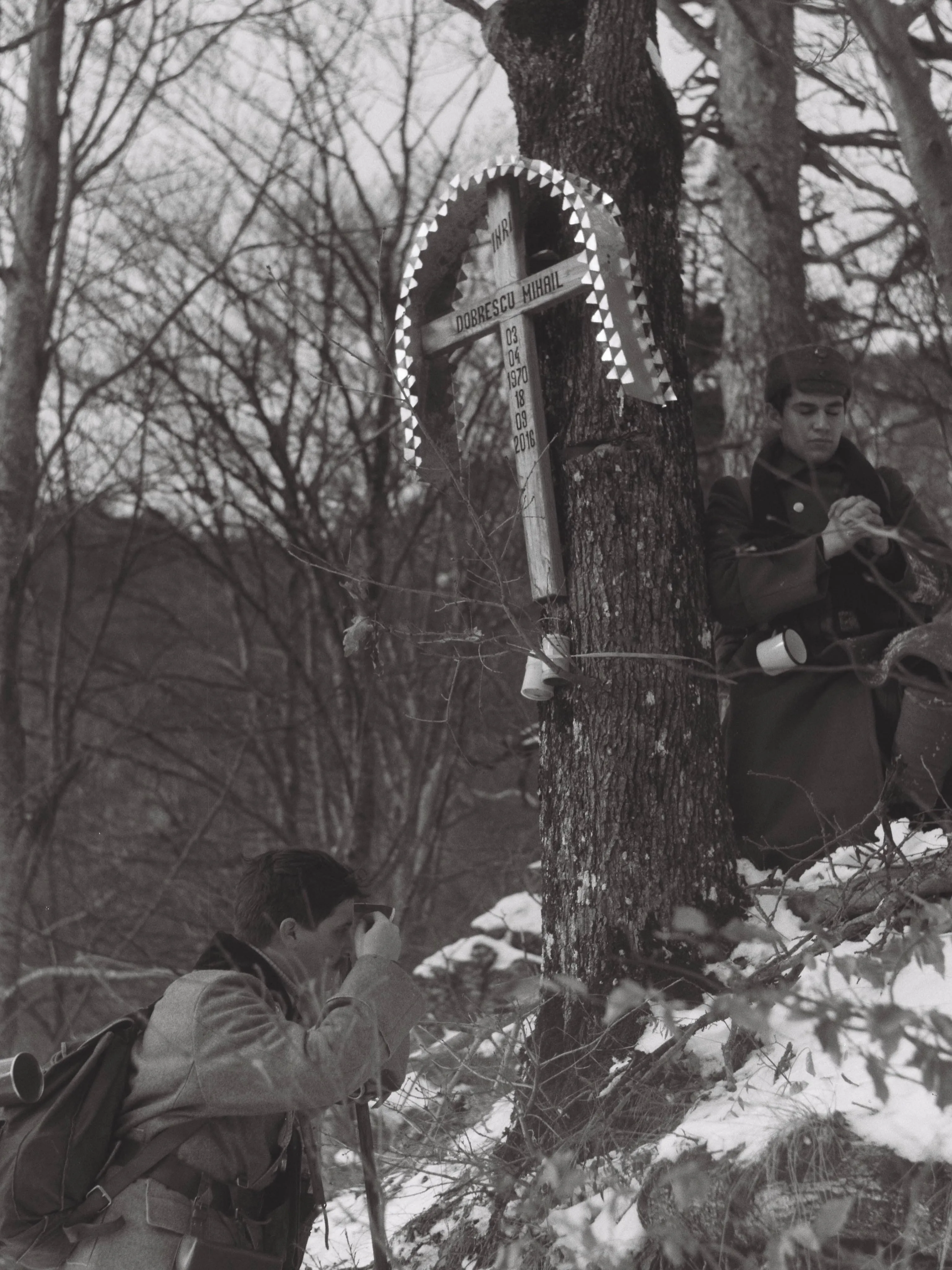 Two people in military uniforms beside a wooden memorial cross in a snowy woodland.